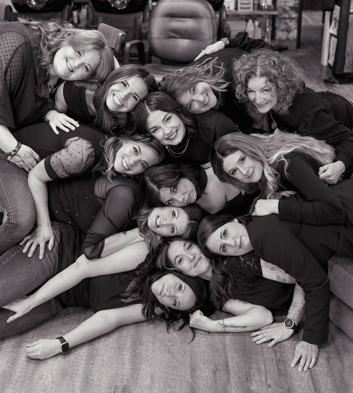 A group of smiling women in black outfits, posing close together on the floor in a joyful manner.